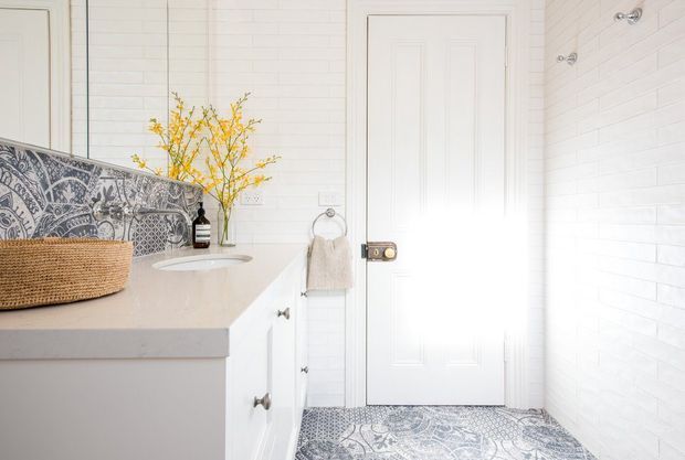 Bright, compact bathroom with white painted brick-style wall tiles, a white paneled door, and a pale stone vanity with integrated basin and white cabinetry. A patterned blue-grey tile backsplash and matching floor create a decorative feature, complemented by a woven basket and a vase of yellow branches.