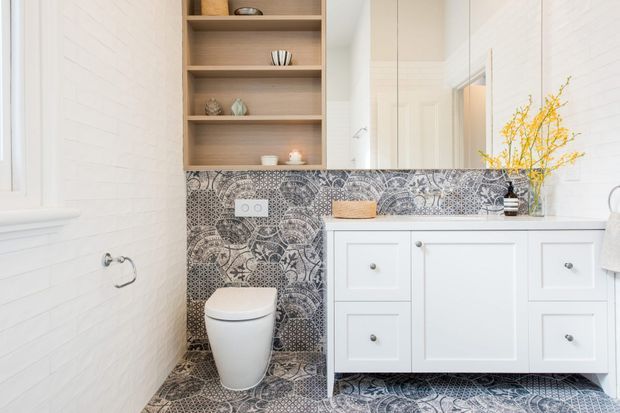 Bright, compact bathroom featuring a white vanity with integrated basin, a contemporary toilet, white painted brick-look wall tiles, and an intricate monochrome patterned tile splashback and floor. Open timber shelving provides display storage above the feature tile, with a mirror and soft decorative styling adding warmth.