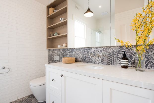 Bright contemporary bathroom with a white vanity, integrated basin, large mirrored cabinet, open timber shelving, white wall tiles, and a patterned decorative tile splashback. A toilet, shower glazing, pendant light, and botanical decor are also visible.