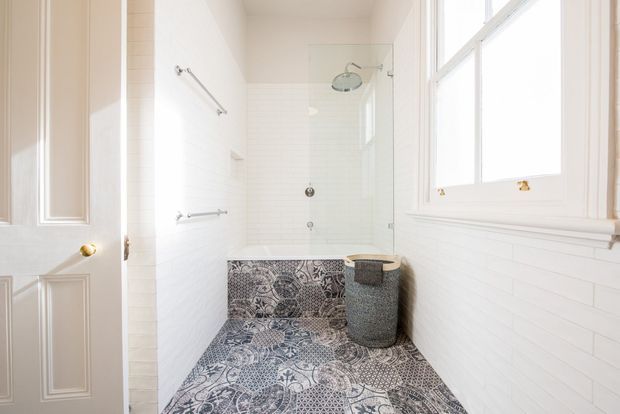 Bright, narrow bathroom with white horizontal wall panelling and a large white-framed window. A glass shower enclosure with a wall-mounted shower occupies the rear, while the floor and lower bath or ledge area feature an ornate monochrome patterned tile. Brass hardware and a grey textured cylindrical stool or hamper add small contrasting details.
