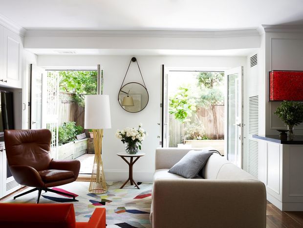 Bright, refined contemporary living room with white panelled walls and cabinetry, timber flooring, large glazed doors opening to a planted courtyard, a neutral sofa, brown leather lounge chair, sculptural floor lamp, patterned rug, and colourful abstract artwork.