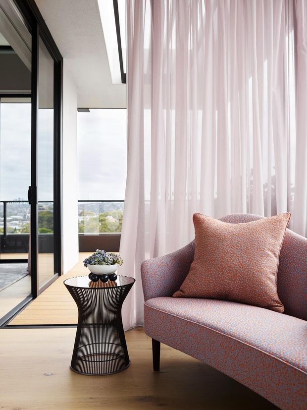 A refined contemporary sitting area with a patterned upholstered armchair, slim black legs, a sculptural black wire side table, and sheer blush-pink curtains. Pale timber flooring, white walls, and black-framed floor-to-ceiling glazing create a bright interior overlooking a distant urban landscape.