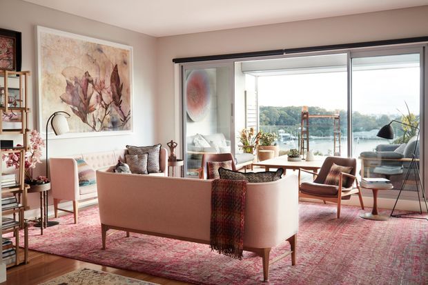 Bright, refined living room with a blush-pink sofa, coordinating armchairs, patterned area rugs, a large floral artwork, open shelving, indoor plants, and floor-to-ceiling sliding glazing overlooking a leafy waterfront setting. The room combines soft contemporary furnishings with warm timber and metal accents.