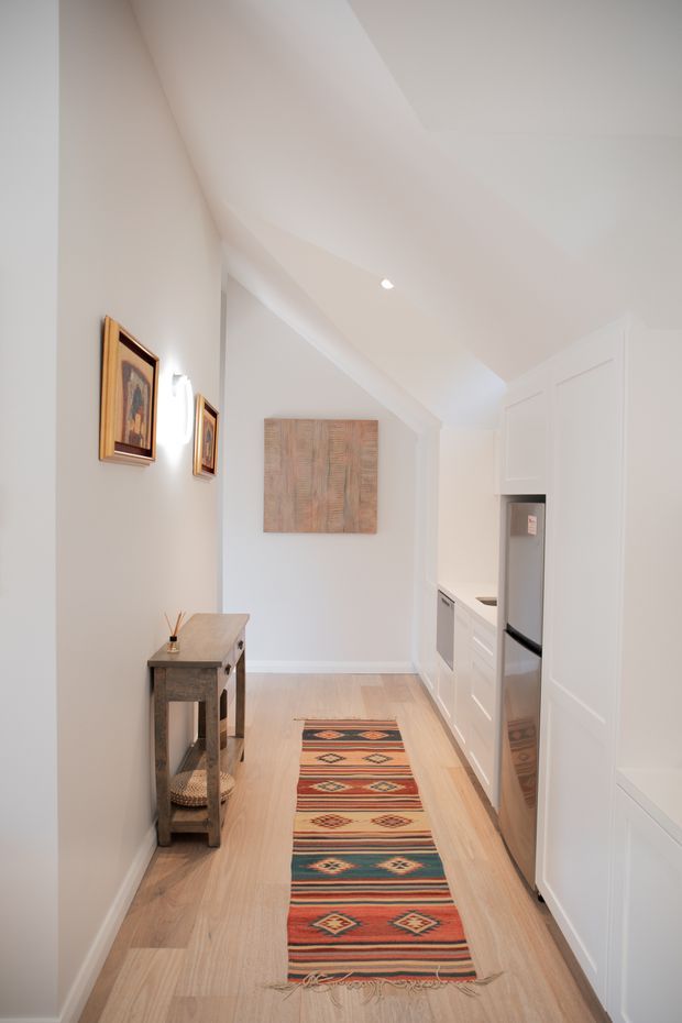A narrow, bright interior corridor with a sloped white ceiling and recessed lighting. White built-in cabinetry and a stainless-steel refrigerator line the right side, suggesting a compact kitchen or kitchenette zone. Pale timber flooring runs throughout, while a slim console table, framed artwork, wall-mounted lighting, and a colourful geometric runner add warmth and character.