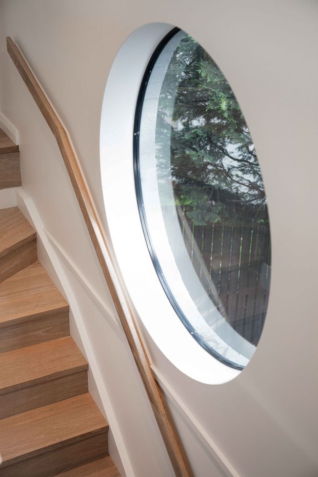 Interior view of a modern stairway with warm timber treads and a slender timber handrail set against white walls. A large oval-shaped window with a white deep reveal and dark frame provides a distinctive architectural feature, looking onto greenery outside.