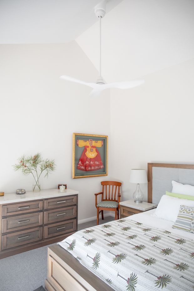 Bright, airy bedroom with white walls and ceiling, a contemporary white ceiling fan, and a large timber-framed upholstered bed. A dark timber dresser with a pale stone-like top sits against the wall, accompanied by a matching bedside table, glass lamp, and vintage wooden armchair. The room includes framed artwork and a light patterned bedspread, creating a clean, warm, mid-century-inspired interior.