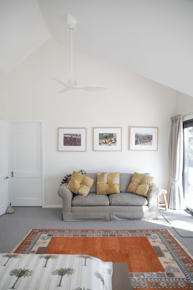 Bright, compact living room with a high sloping ceiling, white walls, a white ceiling fan, grey upholstered sofa, patterned mustard cushions, framed artwork, a large orange and multicolour patterned rug, and pale carpet. A white panel door is on the left and full-height curtained glazing is on the right.
