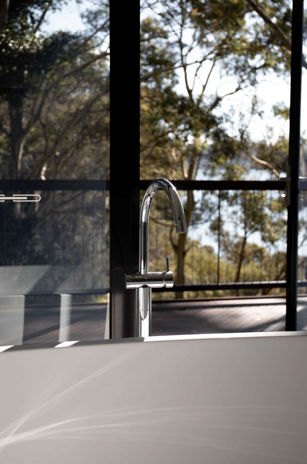 Close-up architectural photograph of a freestanding white bathtub with a polished chrome gooseneck mixer tap, positioned beside full-height black-framed glazing. A leafy treed landscape and timber deck are visible beyond the windows, creating a strong indoor-outdoor connection.