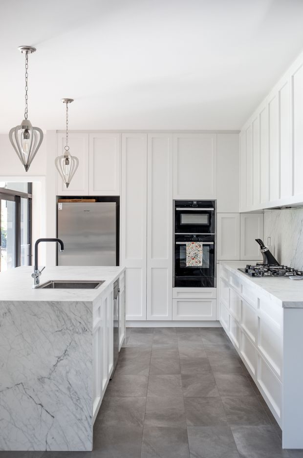 Bright, refined kitchen with extensive white shaker-style cabinetry, a large marble-look waterfall island, integrated stainless-steel refrigerator, built-in black double ovens, gas cooktop, and a pale veined stone backsplash. Two pendant lights hang above the island, while large grey-format floor tiles provide contrast.