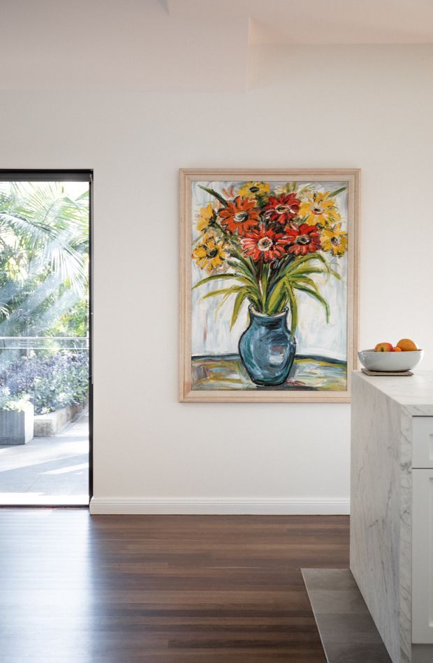 A bright, refined interior view beside a kitchen island, featuring dark timber flooring, pale walls, a large framed floral artwork, and a marble-clad island with white cabinetry. Full-height glazing at left opens toward leafy outdoor planting, bringing in abundant natural light. A bowl of fruit sits on the island, while the composition primarily highlights the wall artwork, flooring, glazing, and kitchen edge.