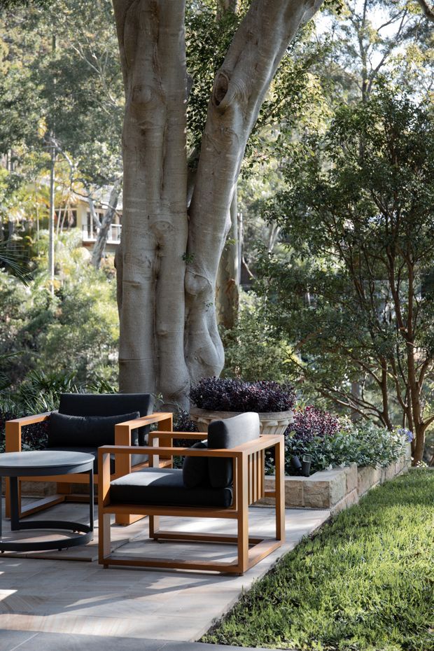 A landscaped outdoor seating area with two black upholstered wooden armchairs and a matching round coffee table arranged on a paved terrace. The space sits beneath mature trees beside raised stone garden beds with dense planting, creating a shaded garden lounge.