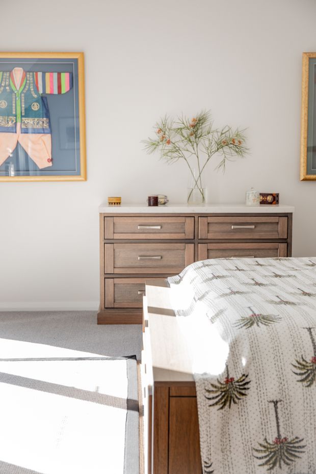 Bright, calm bedroom interior featuring a timber bed in the foreground, a mid-century-style wooden dresser with six drawers against a light neutral wall, framed textile artwork, and a glass vase with delicate branches. Soft grey carpet and strong sunlight create a warm, airy atmosphere.