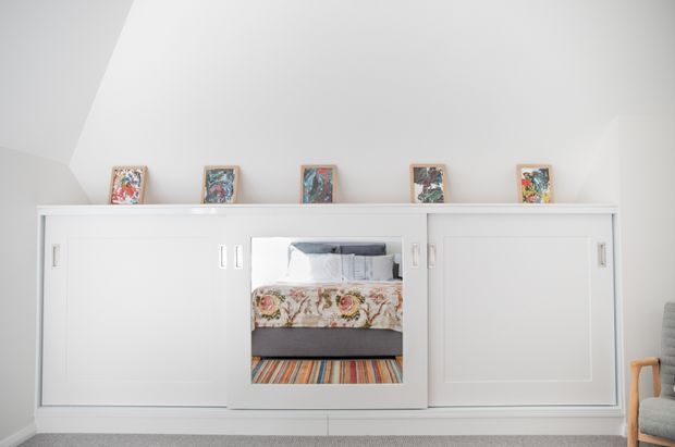 Bright, compact bedroom with a built-in white storage wall featuring sliding-panel doors and a central open niche framing the bed. The bed has floral bedding and a multicoloured striped rug or throw beneath it, while small framed artworks are displayed along the top shelf. A grey upholstered chair is partially visible at the right edge. The space has a clean, restrained built-in design with a sloped or angled ceiling transition.