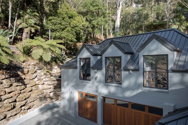 Exterior view of a contemporary multi-gabled building set into a heavily wooded, sloping landscape. The facade combines pale grey rendered walls with dark standing-seam metal roofing, tall dark-framed windows, and warm timber garage or entry doors. A rough stacked-stone retaining wall and native forest planting frame the building.