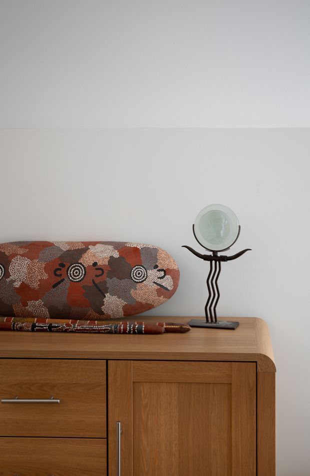 Close-up interior detail of a warm timber sideboard with integrated drawers and cupboard doors, topped with an Aboriginal-inspired patterned textile or artwork and a sculptural decorative object. The composition is minimalist and softly lit, with a plain light neutral wall behind it.