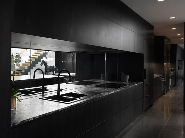 Dark, sophisticated contemporary kitchen with extensive black timber-look cabinetry, a large grey stone island benchtop, integrated cooktop, double black gooseneck sinks and a mirrored splashback. The mirror reflects a sculptural staircase and adjoining dining/living area. Recessed ceiling lights and dark tiled flooring create a dramatic, refined atmosphere.