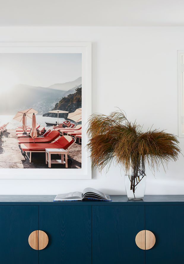 A refined interior vignette featuring a deep blue built-in sideboard with large round light-wood handles, topped with an open book and a glass vase of dried ornamental grasses. Above it hangs a large framed coastal beach photograph, while a white-framed window is visible at right. The palette is crisp white, deep navy-blue, and warm natural brown, with a clean contemporary feel.