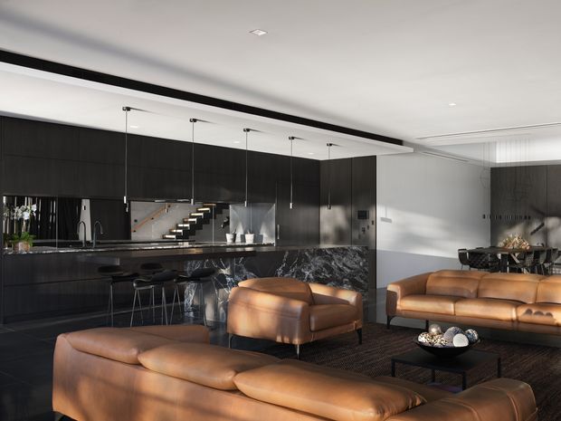 Open-plan contemporary kitchen and living area with dark full-height cabinetry, a marble-patterned island bench, slim black pendant lights, and a leather seating arrangement around a low black coffee table. The space has white ceilings and walls, dark timber or charcoal finishes, warm tan leather furniture, and a staircase visible behind the kitchen.