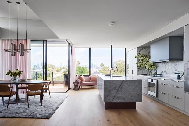 Bright open-plan apartment interior combining a contemporary kitchen, dining area and living space. The room features floor-to-ceiling glazing with city views, pale timber flooring, a large dark stone island, streamlined grey cabinetry, a dining table with mid-century-inspired chairs, a blush sofa, and pendant lighting. The overall look is refined, airy and modern.
