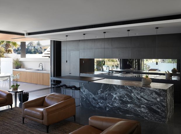 Open-plan contemporary kitchen and living area with dark timber or wood-look cabinetry, a dramatic veined stone island and benchtop, black-framed glazing, and warm brown leather lounge chairs. The kitchen opens to a bright outdoor terrace, with pendant lights, integrated appliances, and reflective splashback or glazing creating a spacious, refined entertaining zone.