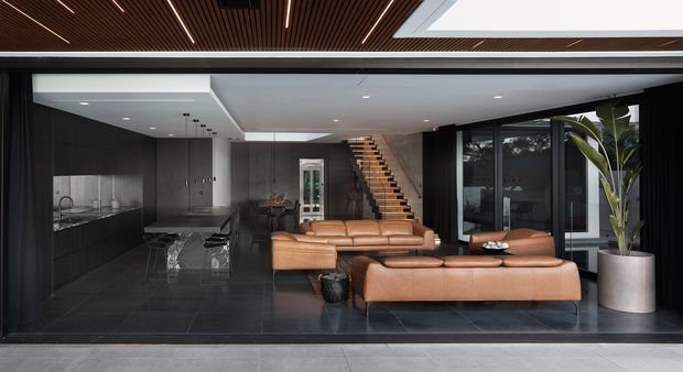 A sophisticated open-plan interior viewed through wide glazing, combining a dark kitchen with island, a warm tan leather lounge arrangement, and a timber staircase. The room features glossy dark tiled flooring, charcoal wall cabinetry, extensive black-framed glazing, recessed lighting, a dramatic slatted timber ceiling, and a large skylight. A tall indoor plant adds greenery near the window.