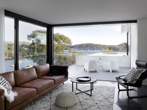 Bright contemporary living room with floor-to-ceiling corner glazing overlooking a leafy waterfront setting. The room features a brown leather sectional sofa, black leather lounge chair, round black coffee table, textured light rug, and a small white outdoor-style seating area on the glazed terrace or balcony beyond.