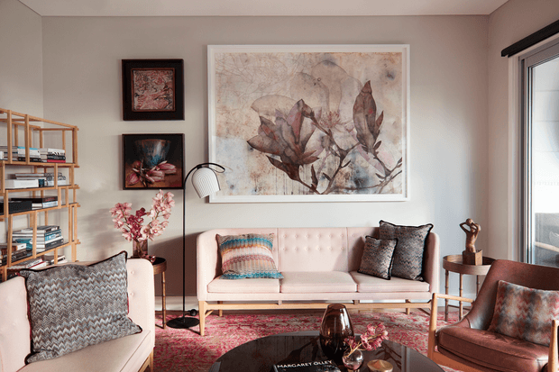 A warmly styled living room with a blush-pink tufted sofa, patterned cushions, a cream armchair, dark wood coffee table, open timber shelving, framed botanical artwork, floor lamp, and richly patterned rug. The room has a refined vintage-inspired scheme with soft neutrals, dusty pinks, terracotta, and dark accents.