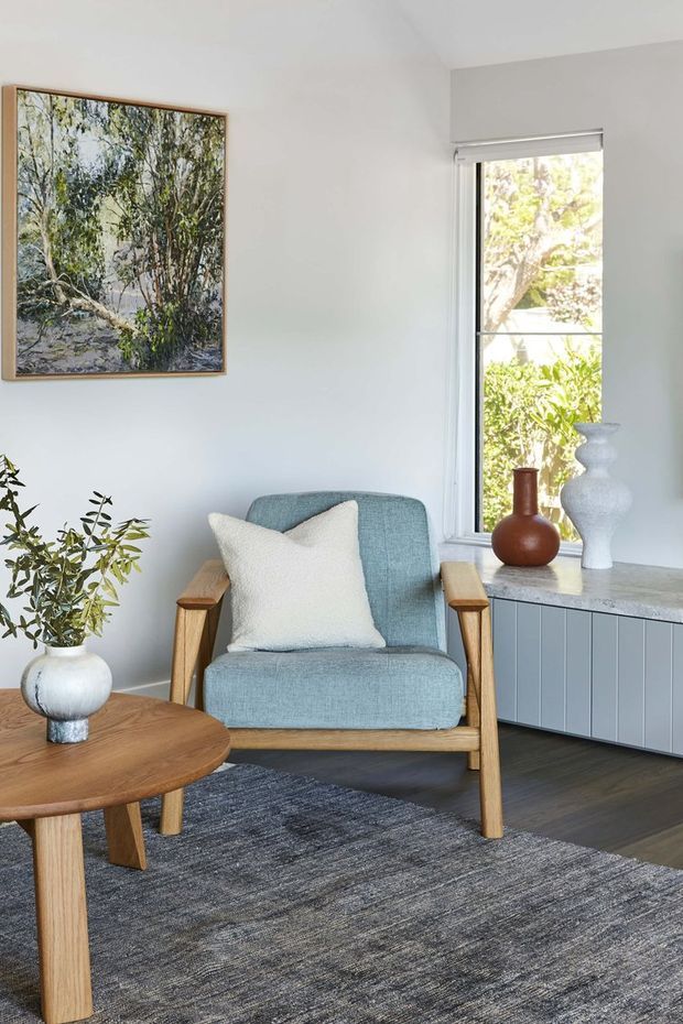 A bright, calm sitting area with a pale blue upholstered armchair, light timber arms and legs, a white cushion, and a round timber coffee table. A large framed nature artwork hangs on the white wall, while a tall window overlooks greenery. A built-in low cabinet with pale blue-grey vertical-panel doors and a light stone benchtop sits beneath the window. A dark grey textured rug and indoor plants complete the space.