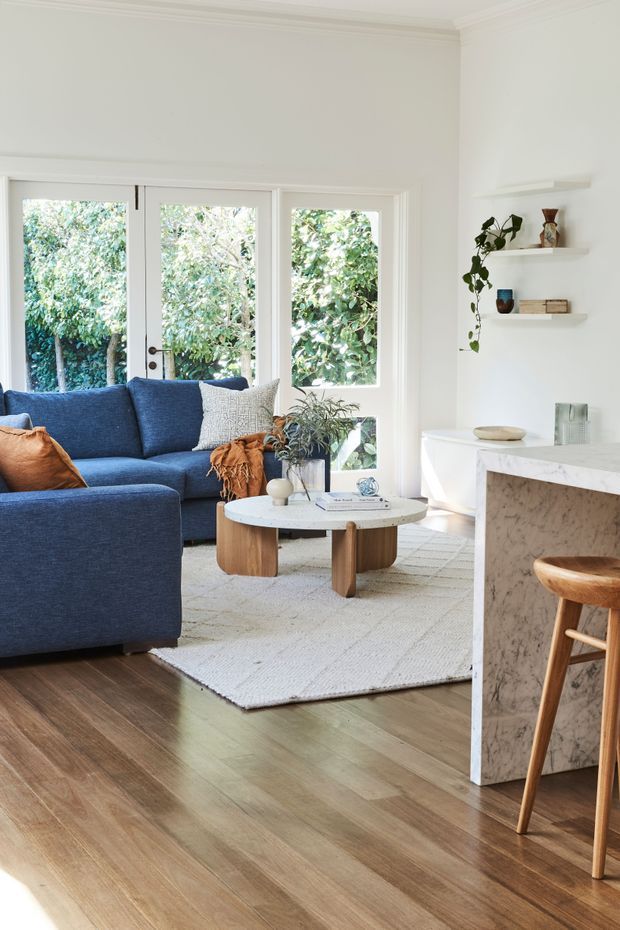 Bright, inviting living room with a deep blue sectional sofa, round timber coffee table, textured light rug, and large white-framed glazed doors opening to leafy greenery. A marble-faced kitchen island with timber stools is visible at right, along with minimalist floating shelves and indoor plants. The space has warm timber flooring, white walls, and abundant natural light.
