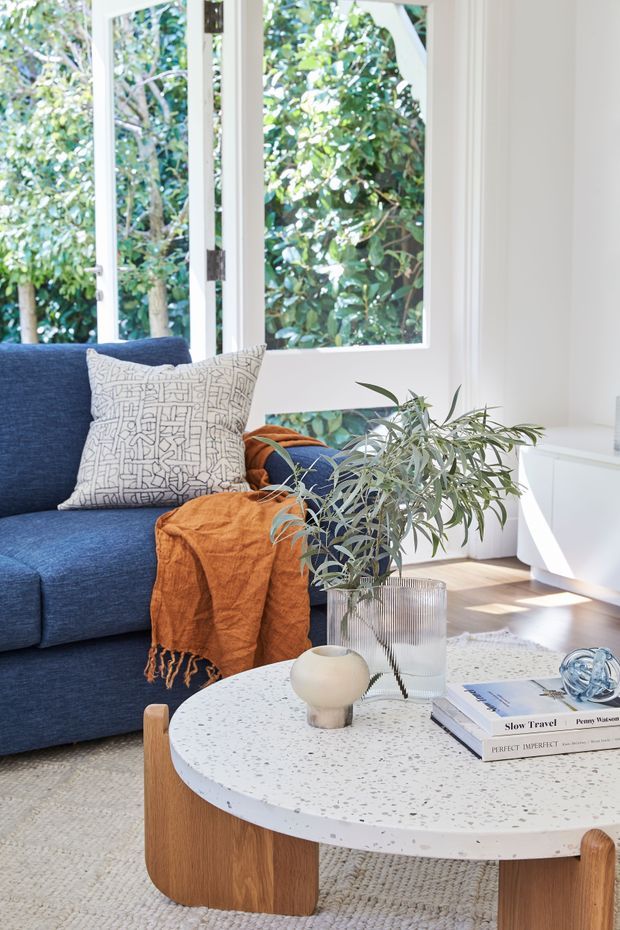 Bright, airy living room with a deep blue sofa, patterned cushion, and rust-orange throw. A round white terrazzo coffee table with a timber base occupies the foreground, styled with a glass vase, greenery, ceramic vessel, books, and a small decorative object. Large white-framed windows overlook leafy greenery, with warm timber flooring and a textured neutral rug.
