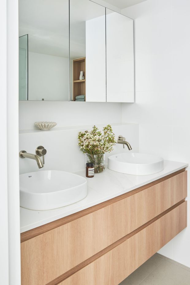 Bright, minimalist bathroom vanity with two white vessel basins, brushed metal wall-mounted taps, a white benchtop, pale timber drawer cabinetry, and mirrored wall cabinets. Open timber shelving adds a small warm accent, while the overall palette is clean white and light wood.