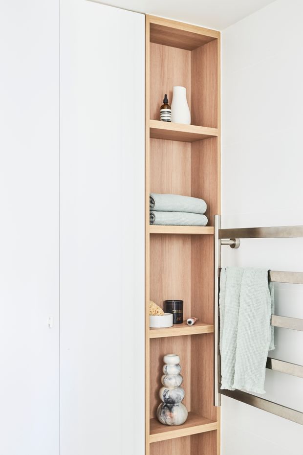 A bright, minimalist bathroom detail showing a tall built-in open timber shelving unit set into a white wall beside a brushed metal heated towel rail. The shelves contain neatly folded towels and small decorative objects, with a pale green towel hanging on the rail. White wall surfaces and clean contemporary joinery create a calm, uncluttered appearance.