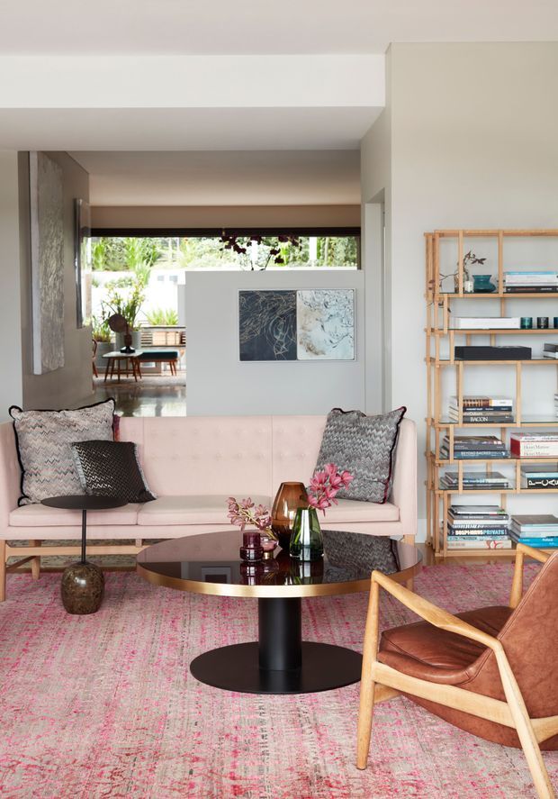 A refined living room with a pale pink upholstered sofa, patterned cushions, a round dark coffee table with a brass-toned rim, and a timber-framed leather armchair. A large patterned rug adds muted pink and grey colour across the floor. Open timber shelving displays books and decorative objects, while a wide opening leads to a bright adjoining indoor-outdoor area. The room has white walls, strong horizontal ceiling beams, and a layered, artful contemporary interior.