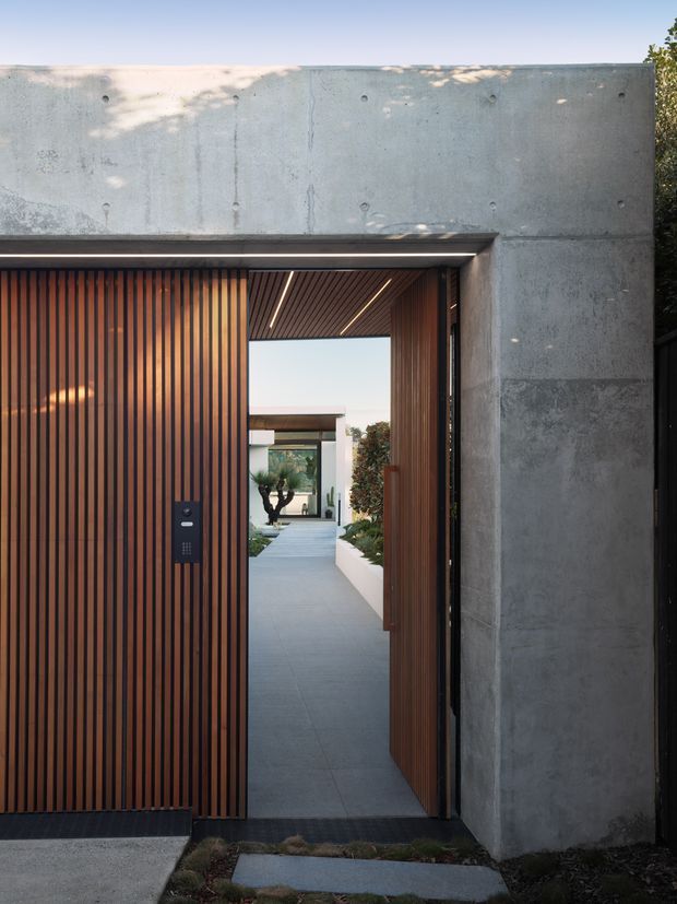 Modern exterior entry viewed from outside, featuring a tall raw concrete wall and a large timber slatted gate and pedestrian door. The open entry reveals a minimalist paved courtyard path, white rendered walls, planting, and a timber-lined soffit with recessed linear lighting.