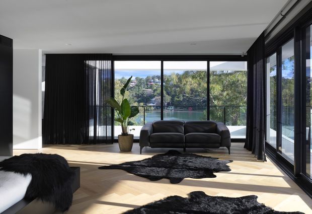 A bright, contemporary living room with pale timber flooring, a dark leather sofa, black curtains, extensive black-framed glazing, and black-and-white hide rugs. The room opens visually to a leafy waterside landscape beyond the glass, with strong natural light and a neutral black-and-white palette.
