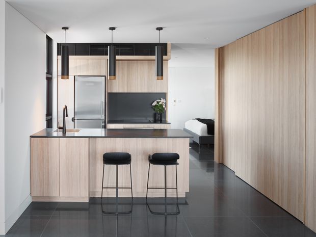 A compact contemporary open-plan kitchen with a pale timber island and full-height timber wall cabinetry, dark stone surfaces, integrated stainless-steel appliances, black pendant lights, and glossy dark flooring. A bedroom is visible beyond the kitchen, creating a streamlined apartment-style living space.