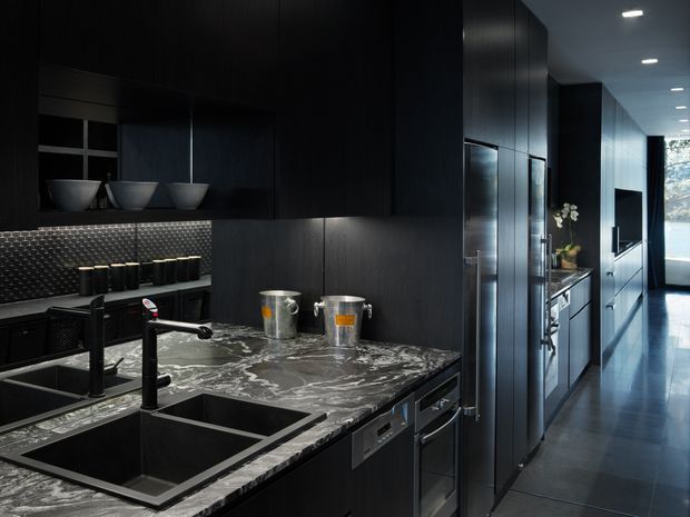 A dramatic, narrow contemporary kitchen with near-black full-height cabinetry, a dark veined stone island benchtop, integrated black double sink and tapware, and stainless-steel appliances. A reflective textured splashback, under-cabinet lighting, and a long corridor-like galley arrangement lead toward bright glazing at the rear.