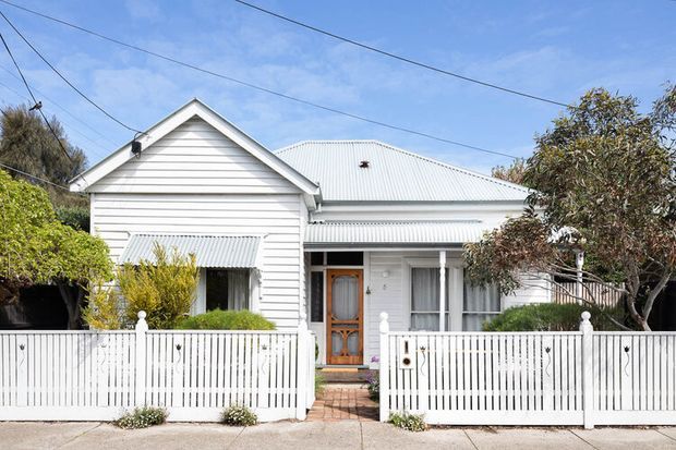 Street-facing view of a traditional single-storey white weatherboard house with a pitched gable roof, corrugated metal roofing, decorative awnings, timber entry door, and a white picket-style front fence with landscaped planting.