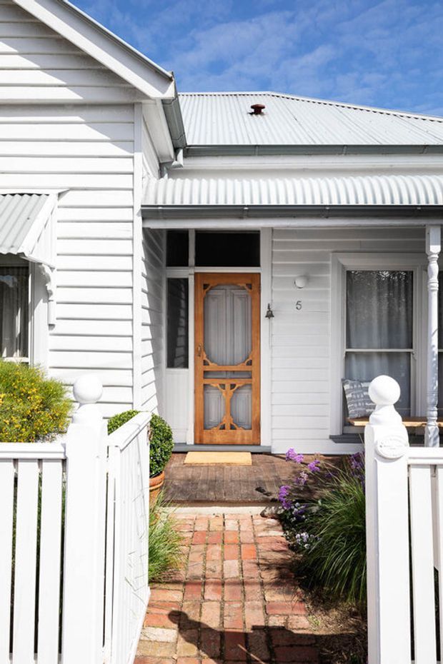 Bright daytime exterior view of a traditional white weatherboard cottage entry. The facade features horizontal timber cladding, a corrugated metal gabled roof and porch awning, a decorative timber front door with glazed panels, turned porch posts, and a small timber deck reached by a red brick garden path. White picket-style fencing and landscaped planting frame the entry.