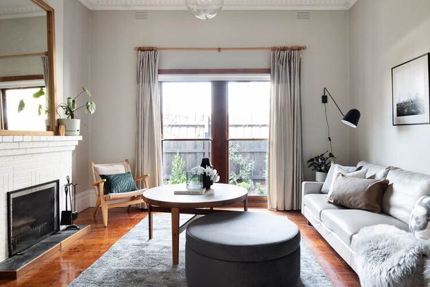 Bright, refined living room with a white-painted brick fireplace, timber flooring, large central rug, round timber coffee table, upholstered ottoman, and a light sofa arranged around the fireplace. Tall glazed timber doors open to a small outdoor area, while grey curtains, a black floor lamp, framed artwork, and classic ceiling cornices add character. The room blends traditional architectural detailing with contemporary furnishings.