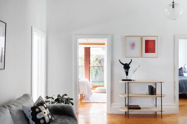 Bright, refined living room with white walls, timber floors, a grey sofa, indoor plants, framed artwork, and an open black metal-and-timber console. Doorways lead to a softly furnished room and a bedroom, while a glass pendant light hangs overhead.