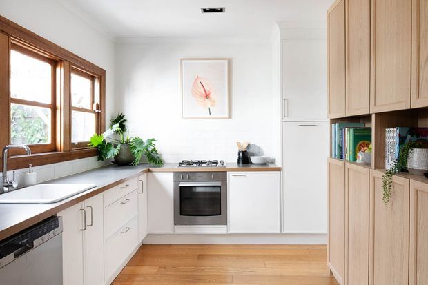Bright, compact L-shaped kitchen with white shaker-style cabinetry, integrated appliances, a timber benchtop, warm timber flooring, and a large built-in timber display and storage unit. Natural light enters through timber-framed windows above the sink, with indoor plants and understated decor adding a fresh, lived-in feel.