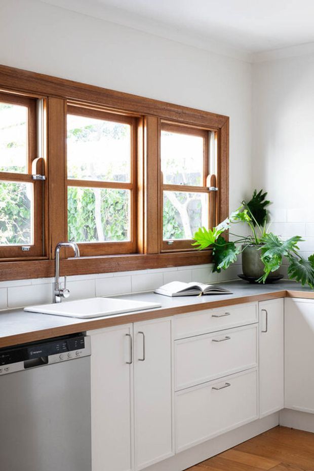 Bright, compact kitchen with white shaker-style cabinetry, a pale stone benchtop, integrated sink and chrome tap, stainless-steel dishwasher, and a white tiled splashback. Warm timber-framed windows dominate the wall above the counter, with potted greenery and an open book adding natural accents. Timber flooring and abundant daylight create a clean, welcoming atmosphere.