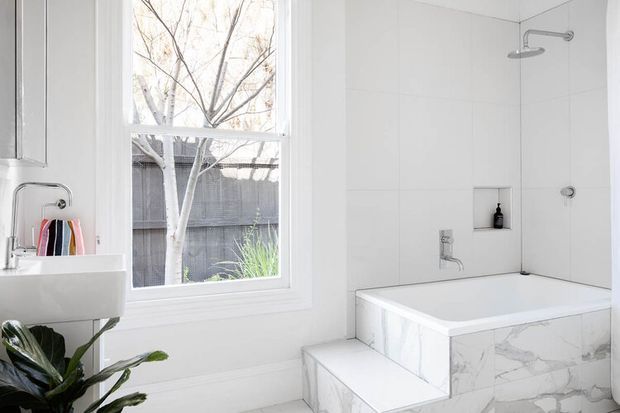 Bright contemporary bathroom featuring a white wall-mounted basin, frameless mirrored cabinet, large white tiled walls, a walk-in shower with overhead and handheld fittings, and a built-in bathtub clad in white marble-look tile. A large window provides abundant natural light and overlooks a fenced garden.