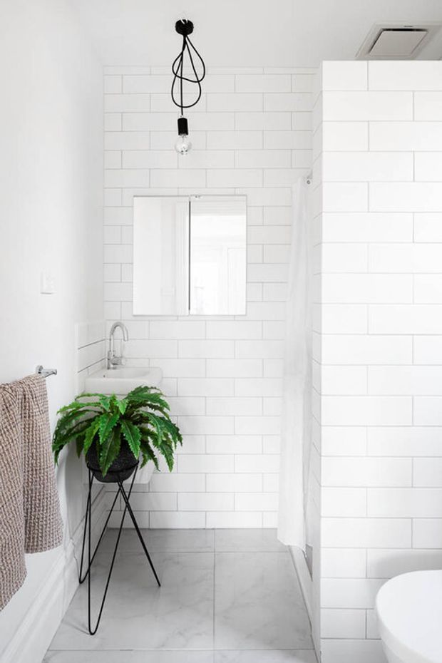 Bright, compact modern bathroom with white subway-tile walls, pale grey tiled flooring, a wall-mounted mirror, small white basin with chrome tapware, and a freestanding bathtub partially visible behind a tiled partition. A black pendant light and leafy plant add contrast and texture.