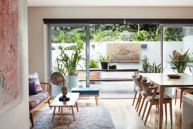 Bright open-plan living and dining area with full-height sliding glass doors opening to a landscaped courtyard. The room features pale timber flooring, a light neutral palette, mid-century-inspired timber furniture, a colourful patterned armchair, indoor plants, and a large dining table. A sculptural textured wall artwork and built-in outdoor bench are visible beyond the glazing.