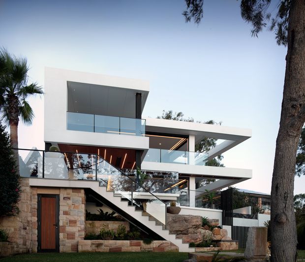 Exterior view of a contemporary multi-level residence with strong horizontal cantilevered forms, extensive floor-to-ceiling glazing, glass balustrades, a broad external stair, exposed stonework at the lower level, and integrated warm linear lighting beneath the projecting slabs. Mature trees and landscaped planting surround the house.