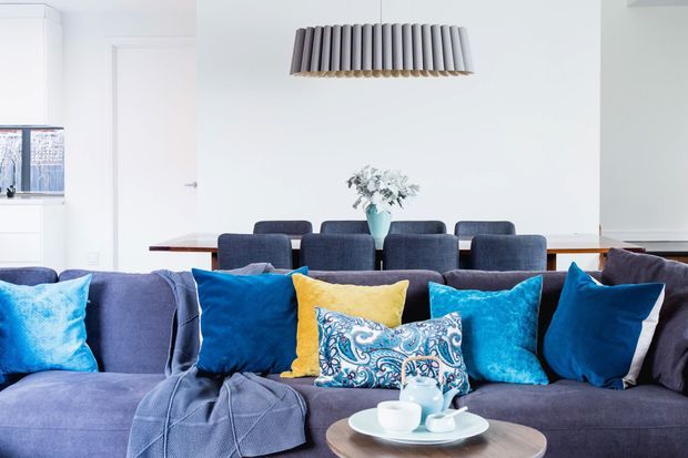 Bright, open-plan living and dining area with a large blue upholstered sectional sofa in the foreground, layered blue, turquoise, yellow and patterned cushions, and a round timber coffee table with a tea set. Behind it is a dining table with dark upholstered chairs, white walls and cabinetry, and a distinctive suspended ribbed pendant light.