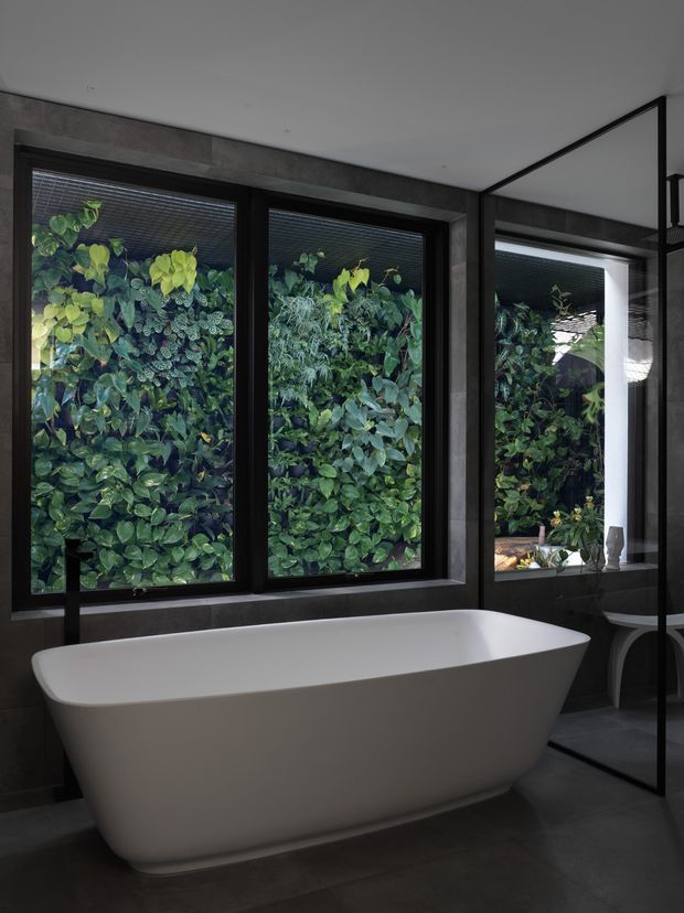 A dark, contemporary bathroom featuring a freestanding white sculptural bathtub positioned beneath large black-framed windows. Dense green foliage is visible directly outside, while dark grey tiled walls and flooring create a dramatic, refined setting. A glass shower enclosure and partial vanity area are visible to the right.