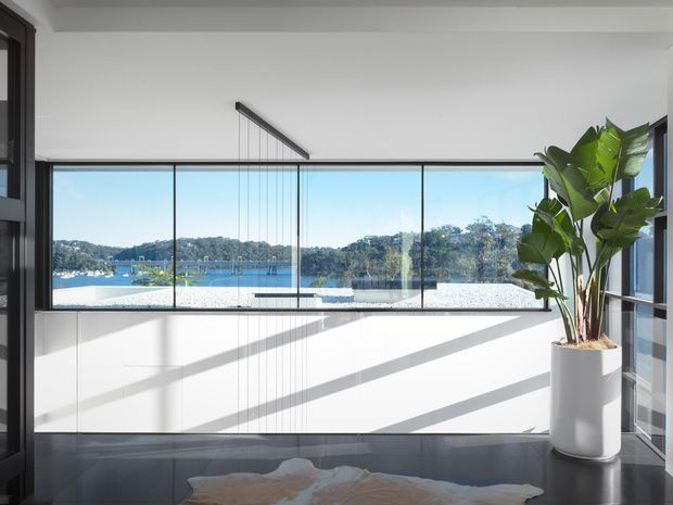 Bright contemporary interior with floor-to-ceiling glazing overlooking a calm waterfront setting. A large white built-in cabinet spans the window wall, complemented by a dark polished floor, sculptural linear pendant light, and a tall potted tropical plant. Strong sunlight creates dramatic diagonal shadows across the cabinetry and floor.